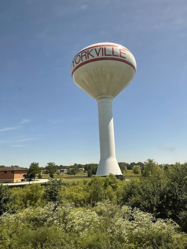 The City of Yorkville water tower, white with red lettering, seen from a passing train in summer